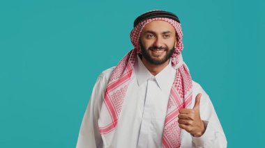 Kendine güveni tam bir adam stüdyoda kendini gösteriyor. Geleneksel Arap kültürünü temsil ederken onaylıyor ve onaylıyor. Orta Doğulu pozitif adam İslami kostüm ve türban takıyor..