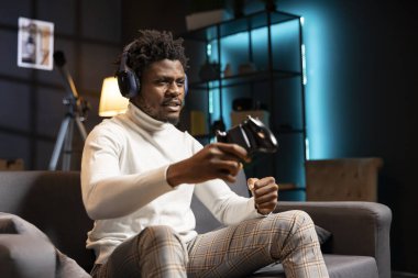 Cyan neon ışıklı apartman dairesinde takım arkadaşlarıyla oyun konsolunda oyun oynayan bir adam. BIPOC oyuncusu esport oyununda rakiplerle kulaklık kullanarak savaşıyor, arkadaşlarıyla konuşuyor