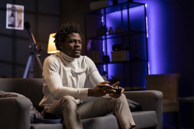 Oyuncu evde oyun oynuyor ve boş zamanların tadını çıkarıyor. Mavi neon ışıklı oturma odasında kanepede oturan Afrikalı Amerikalı adam, konsolda video oyunu oynuyor, yoğun bir iş gününden sonra dinleniyor.