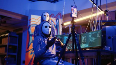 Gizli yeraltı üssünde fidye videosu çeken isimsiz şeytani geliştiriciler, ödeme talepleri karşılanmazsa çalınan verileri halka açıklamakla tehdit ediyorlar, cep telefonuyla şantaj görüntülerini kaydediyorlar.