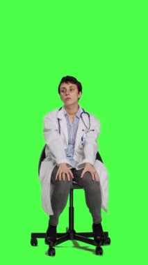 Öndeki beyaz önlüklü doktor hastaları muayenede bekliyor, yeşil perde arkaplanına karşı bir sandalyede oturmak için sabırsızlanıyor. Steteskoplu tıbbi pratisyen insanları bekliyor.