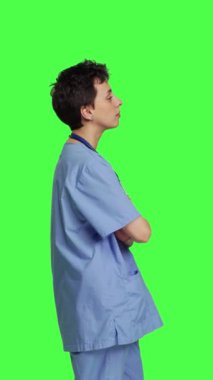 Gülümseyen tıbbi asistanın portresi kollarını kavuşturup mavi hastane önlüğü giymiş bir şekilde poz veriyor. Yeşil perde arkaplanına karşı başarılı bir hemşire, sağlık uzmanı.