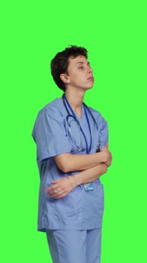 Klinikte hastaları beklerken sabırsız hemşire sinirlenmiş gibi davranıyor, yeşil ekran arkaplanının karşısında duruyor. Endişeli mavi önlüklü hemşire insanların kontrole gelmesini bekliyor.