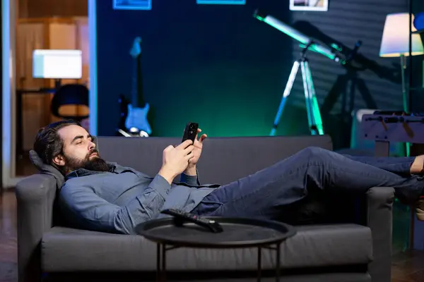 冷冰冰的男人在家里悠闲地躺在沙发上 用智能手机给朋友们发短信 躺下的人 在客厅里用手机在网上和同伴聊天 — 图库照片