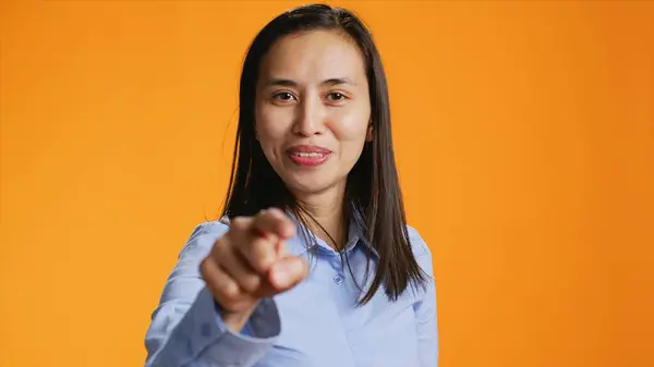 Happy asian person pointing finger at camera during shoot time. Positive young woman with casual attitude using charisma, standing in studio over orange background, showing something.