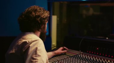Müzik yapımcısı kabloyu amfi ya da ses kompresörünün içinde tamir ediyor, kontrol odası teknik ekipmanlarını kayıt seansı için hazırlıyor. Ses mühendisi şarkılar üzerinde çalışıyor. Kamera B.