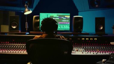 Müzik yapımcısı kontrol odasında kayıt ve düzenleme yapıyor, prodüksiyon sonrası ses efektleri yaratmak için düğmelere basıyor. Ses teknisyeni profesyonel stüdyo ekipmanlarıyla çalışır. Kamera B.