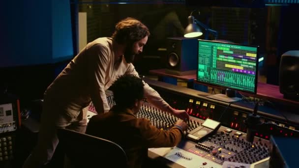 ミュージシャンとオーディオ技術者の多様なチームがヒット曲を作成し プロの音楽スタジオで働いています コンソールパネルをミキシングするプロデューサーの編集と録音トラックを持つアーティスト カメラ — ストック動画
