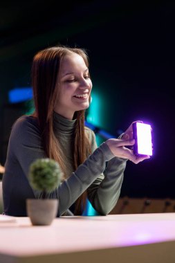 Happy young influencer in studio using camera to capture live broadcast, reviewing mini RGB lights. Joyful media star films electronics haul, presenting wireless LED lighting device clipart