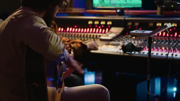 プロのスタジオ コントロール ルームでギターを歌うクリエイティブなミュージシャンが プロデューサーとのトラックを録音した 彼のアルバムのために新しい曲を作曲するシンガーソングライター コンソール上の曲を編集 カメラ — ストック動画