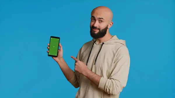 グリーンスクリーンディスプレイでスマートフォンを保持しているムスリム男性は 携帯電話アプリで単離された染色体テンプレートを指摘します 白いコピースペースのモックアップレイアウトを提示する若い大人 カメラ — ストック写真