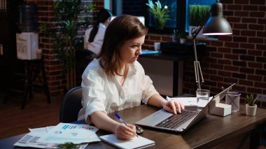 Worker writing financial business details on notepad, crosschecking with information on laptop. Businesswoman in brick wall office focused on finishing company project, camera A clipart