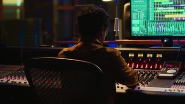 Afrika kökenli Amerikalı müzik yapımcısı profesyonel stüdyodaki yeni şarkısında kontrol odasında müzik karıştırıyor ve ustalaştırıyor. Teknisyen ses panelindeki düğmelere ve sürgülere basıyor. Kamera A.