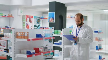 Pharmacy worker writing every medical product on inventory, creating long list with all pharmaceutics and medicaments in drugstore. Pharmacist ensuring variety of healthcare supplies. Camera 1. clipart