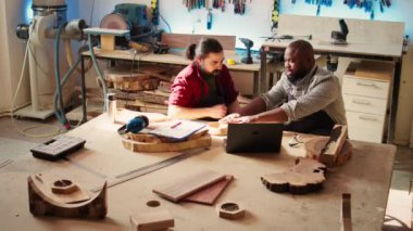 Marangoz ve meslektaşı, tamamlanmış ahşap sanat ürünlerini tasdik ederken, planlarla karşılaştırdılar. Zanaatkar ve çırak, dizüstü bilgisayar ekranı B kamerasındaki şemalara dayalı hasarlar için ahşap nesneyi teftiş ediyor.