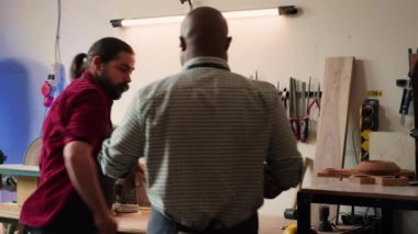 Marangoz, Afrika kökenli Amerikalı iş arkadaşının yardımıyla ahşap malzemeler üzerinde kullanmak için raftan araç seçiyor. Craftsperson, BIPOC çırağı ve kamera A ile kereste parçaları üzerine kalite güvencesi veriyor.