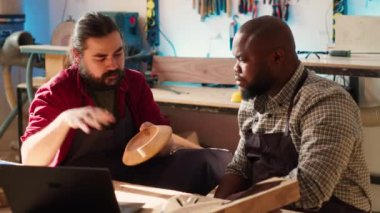 Ahşap işçisi dizüstü bilgisayardaki planları inceliyor, Afrikalı Amerikalı çırak ile beyin fırtınası yapıyor. Üreticilerden oluşan bir ekip, mobilya montaj şemalarını stüdyoda, kamera A 'da tartışıyor.