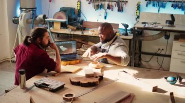 Carpenter, CAD yazılımında çırağıyla birlikte dizüstü bilgisayardaki planları inceliyor. Tahta işçileri stüdyoda defter üzerine özel 3D mobilyalar tasarlıyorlar, müşteri siparişi için hazırlanıyorlar, kamera B