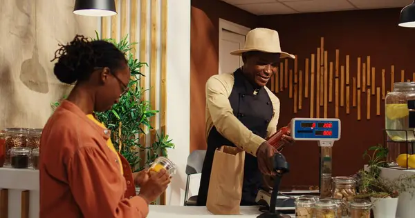 stock image African american woman bringing natural products at checkout, talking to vendor about healthy eating while he scans goods. Local farmer selling his homegrown veggies and homemade items. Camera 2.