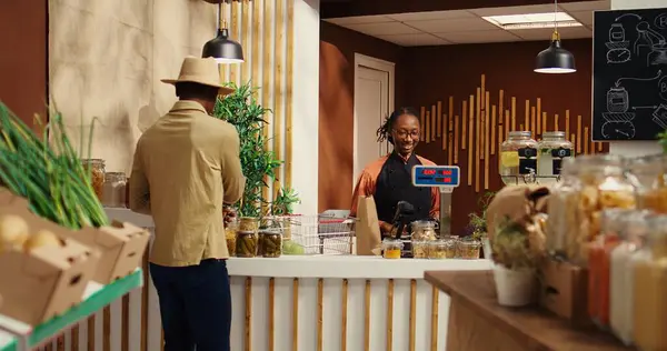 stock image African american seller working at supermarket cash register counter, weighting organic additives free fruits and vegetables. Vendor selling fresh bio produce to male customer at checkout. Camera 2.