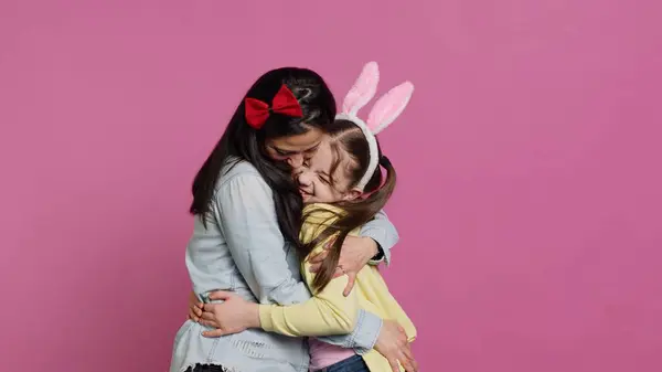 Lovely schoolgirl with bunny ears an her mom waving on camera, having fun and laughing against pink background. Cheerful mother and her daughter embracing and kissing each other. Camera A.