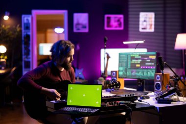 Laptop 'ta yeşil ekran planının yanında akustik gitar çalan bir müzisyen, karıştırma konsolu ve daw yazılımı kullanarak melodileri kaydediyor. Enstrüman ve müzik endüstrisinde yeni anlaşmalar deneyen sanatçı.