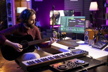 Genç şarkıcı akustik gitar çalıyor ve bilgisayar üzerinde daw yazılım arayüzü kullanarak ev stüdyosunda elektronik aletlerle müzik yapıyor. Ses yapımcısı stereo konsoldaki melodileri ve karıştırma seslerini kaydediyor.