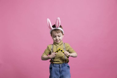 Kameranın önünde renkli bir oyuncakla oynayan neşeli küçük bir çocuk pembe arka planda boş zamanların tadını çıkarıyor. Tavşan kulağı takan ve bir Paskalya fotoğrafçısına poz veren neşeli genç bir çocuk..