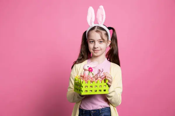 卵とイースターのお祭りのためのウサギで飾られたハンドメイドのバスケットを示す若い楽観的な少女は ピンクの背景の上にポーズ 春の装飾を提示するバニーの耳を持つ小さなかわいい子供 — ストック写真