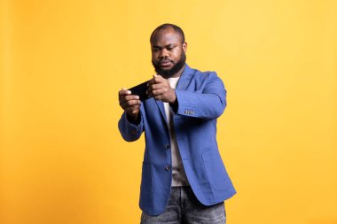 BIPOC çalışanı peyzaj modunda cep telefonuyla video oyunu oynuyor, boş zamanların tadını çıkarıyor. Oyuncu cep telefonuyla eğleniyor, eğleniyor, sarı stüdyo arka planında izole edilmiş.