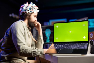 Man with EEG headset on writing code to transfer mind into virtual world, isolated screen laptop on desk Transhumanist using neuroscientific tech and chroma key notebook to gain superintelligence clipart