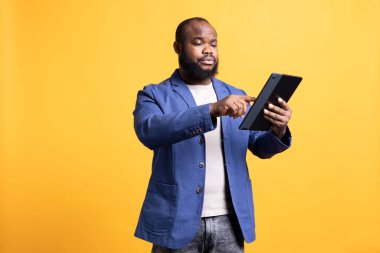 Sıkılmış insan, düşüncesizce tablet ekranda kayıyor, stüdyo arka planında izole edilmiş. Afro-Amerikalı bir adam taşınabilir cihazla sosyal medyada vakit geçiriyor, boş zamanların tadını çıkarıyor.