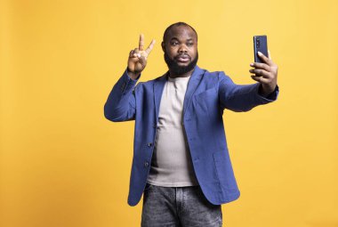 Akıllı telefonu selfie çekmek için kullanan, zafer işareti yapan bir adam. Afro-amerikan kişi fotoğraf çekiyor, cep telefonu ile, barış sembolü jestleri gösteriyor, stüdyo arka planı