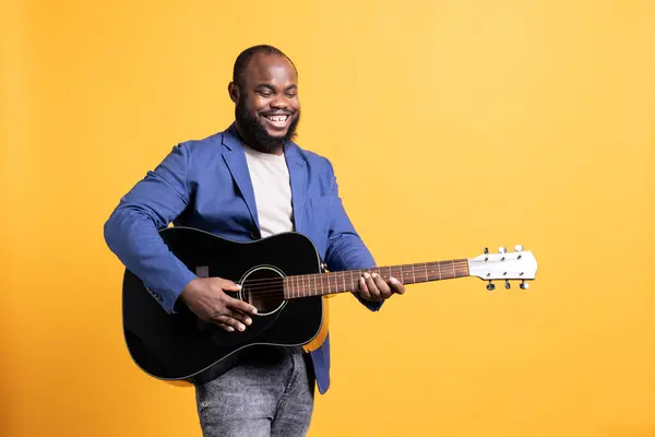 Smiling singer holding guitar, performing blues tunes during concert isolated over yellow studio background. Merry musician performing composition on stringed musical instrument