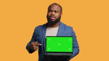 Yeşil ekran dizüstü bilgisayar tutan neşeli Afro-Amerikan adam tavsiye veriyor. Dost canlısı BIPOC çalışanı, model defteri işaret ederek, olumlu geri bildirim, stüdyo arka planı, kamera A