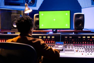 Afrika kökenli Amerikalı ses mühendisi profesyonel kayıt stüdyosunda yeşil ekranla çalışıyor. Paneldeki sesleri karıştırıp ustalaştırıyor. Genç müzik yapımcısı düğmeleri ve faders iterek.