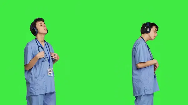 愉快的护士带着无线耳机唱歌跳舞 在绿屏的背景下听着现代歌曲 专业欣赏喜爱的音乐和创作舞步 摄像头B 免版税图库照片