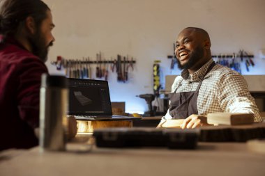 Neşeli çırağı olan mutlu marangoz CAD yazılımında dizüstü bilgisayardaki planları inceliyor. Gülümseyen ahşap işçisi ve neşeli iş arkadaşı deftere özel 3D mobilya dizayn ederken eğleniyorlar.