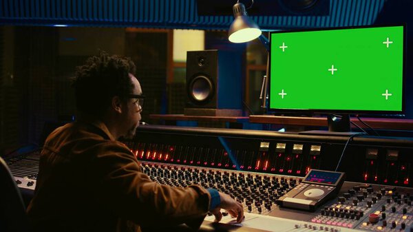African american sound designer looks at greenscreen to record and edit tunes, uses mixing console to create new songs. Skilled technician works on processing and track recording. Camera B.