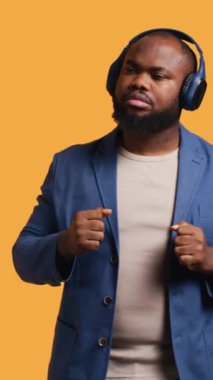Dikey video Mutlu Afro-Amerikan kişi kulaklıkla elektronik şarkılar dinleyip eğleniyor. Neşeli BIPOC DJ gibi davranıyor, dans müziğinden hoşlanıyor, stüdyo arka planı, kamera B