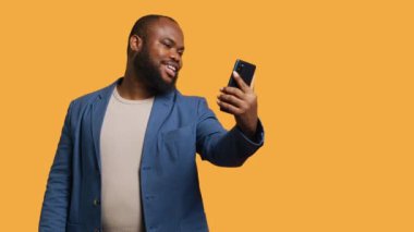 Her açıdan selfie çekmek için cep telefonu kullanan Afrikalı narsist bir adam. Sosyal medya kullanıcısı telefon kamerasıyla fotoğraf çekiyor, mutlu bir şekilde gülümsüyor, stüdyo arka planı, kamera B