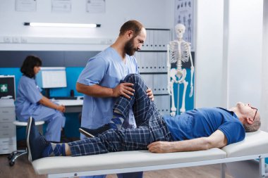 Fizyoterapist, kinesiyoterapi kliniğinde bacağından yaralanan yaşlı bir adama yardım ediyor. Pratisyen hemşire, diz ağrısı ve kas basıncı olan emekli hastaya destek veriyor..