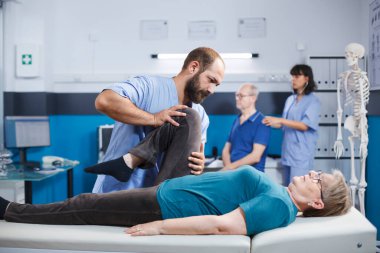 Kayropraktik uzmanı, diz sakatlığı olan yaşlı bir hastaya fiziksel tedavi için yardım ediyor. Osteopatik tedavi ve tedavi için tıbbi asistanın yardımıyla bacak egzersizi yapan kadın..