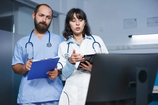 一个拿着剪贴板的白人男人和一个在诊所拿着数码平板电脑的女人谈论病人的预约 女医生与男护士讨论保健和药物治疗 — 图库照片