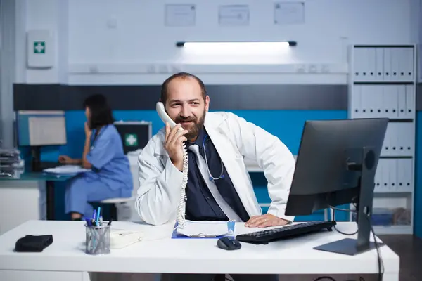 코카서스 의사는 사무실 책상에 전문가와 전화로 이야기 실험실 코트를 남자는 스톡 사진