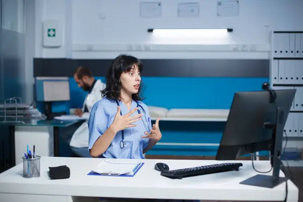 病院の女性看護師は デスクトップPcを使ってビデオ通話をする クリニックのオフィスに座った白人女性がバーチャルミーティングを行った ヘルスケアにおけるコラボレーションと技術 ストックフォト