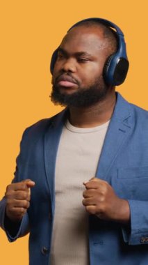 Dikey video Mutlu Afro-Amerikan kişi kulaklıkla elektronik şarkılar dinleyip eğleniyor. Neşeli BIPOC DJ gibi davranıyor, dans müziğinden hoşlanıyor, stüdyo geçmişi, kamera A