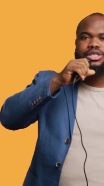 Dikey video Afro-Amerikan şarkıcı mikrofon tutuyor, şarkılar söylüyor, sarı stüdyo arka planında izole edilmiş. Enerjik müzisyen hayali kalabalığa şarkı söylüyor, karaoke yapıyor, kamera A