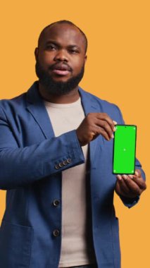 Dikey video Neşeli Afro-Amerikalı adam yeşil ekran akıllı telefon öneriyor, gülüyor, stüdyo geçmişi var. Neşeli BIPOC çalışanı model cep telefonu sunuyor, olumlu geri bildirim veriyor, kamera B