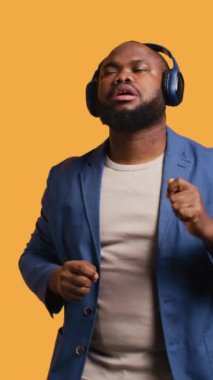 Dikey video Mutlu Afro-Amerikan insan eğleniyor, ritim içinde dans ediyor, kulaklık dinliyor. Neşeli BIPOC adamı kendini eğlendirirken müzik, stüdyo arka planı, kamera B